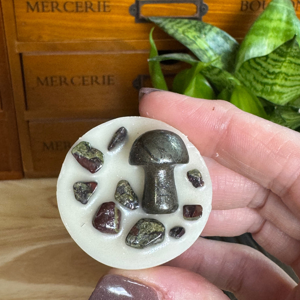 A hand holds a circular white crystal cluster featuring a metallic silver mushroom cabochon and several small polished stones, with a wooden drawer and green plant in the background.