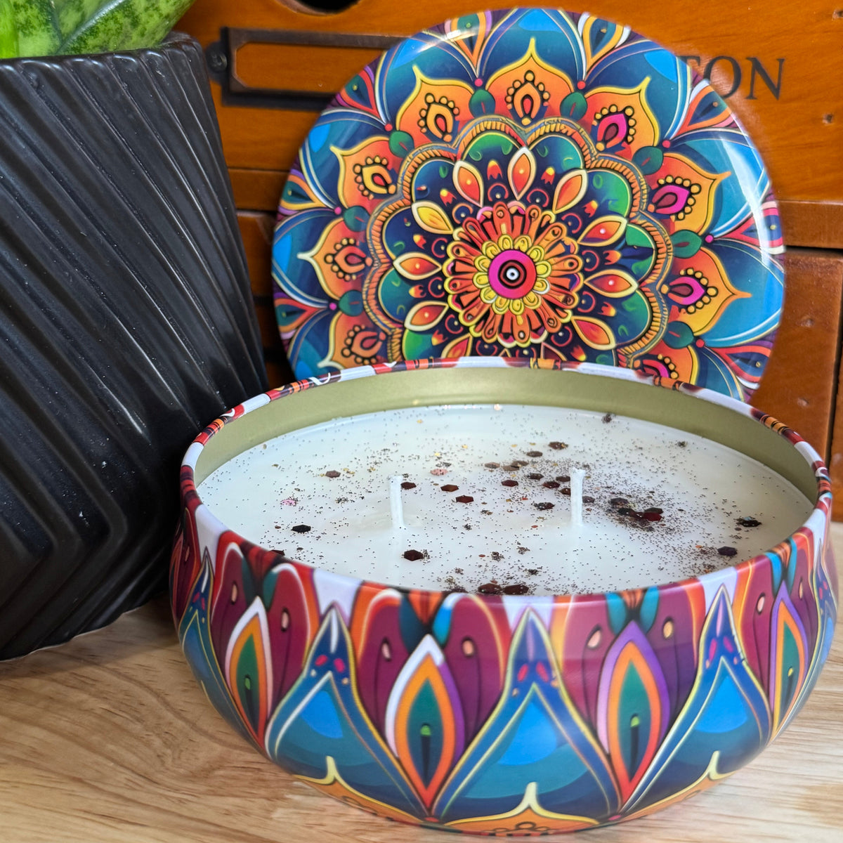 A round metal candle tin with a colorful mandala and geometric pattern, shown open with white wax inside and its matching lid leaning behind it on a wooden surface.