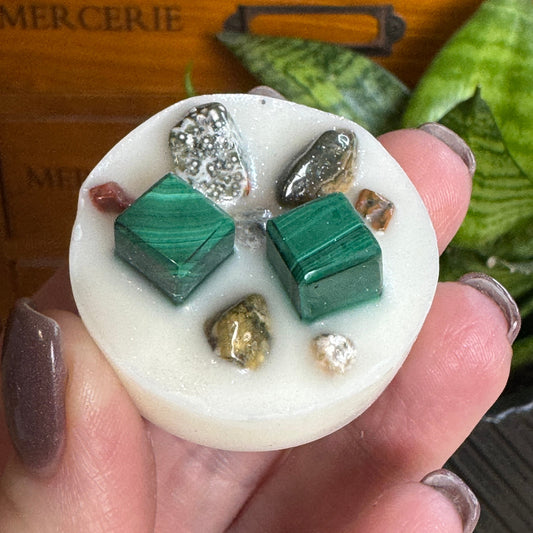 A hand holding a round white wax melt or soap piece decorated with two green malachite cubes, a silver crystal, and small natural stones, with a wooden drawer and green leaves in the background.
