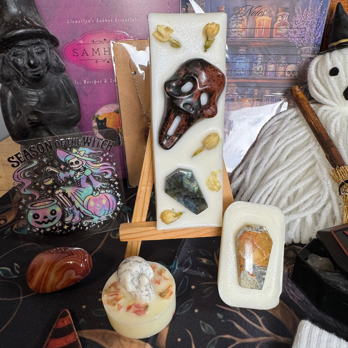 A styled flat-lay of handmade wax melts and crystals, featuring a tall rectangular bar with a skull and dried botanicals on a small wooden easel, a small oval wax melt with a crystal, a round cream candle, and witch-themed props including a black witch figurine, a 'Season of the Witch' card, and a white ghost doll.