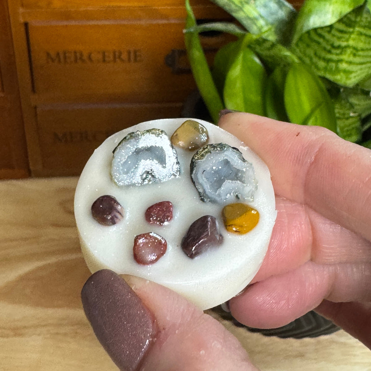 A hand holds a round white soap or candle featuring embedded agate and polished stone pieces, with a wooden drawer and green plant in the background.