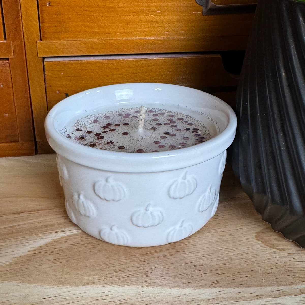 A white ceramic candle holder with a pumpkin pattern, filled with a candle, sitting on a wooden surface in front of a wooden cabinet.