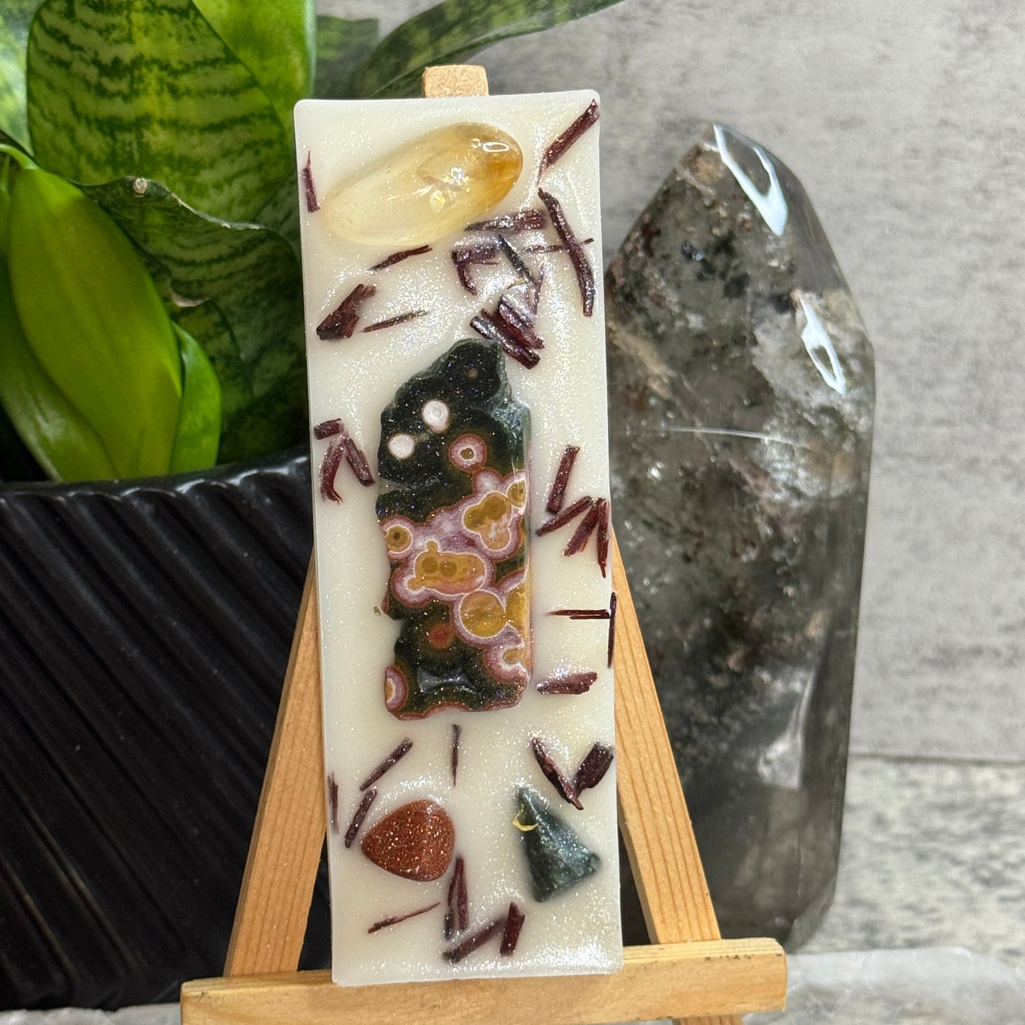 A rectangular cream-colored resin candle displayed on a small wooden easel. The candle features inclusions such as a yellow crystal, a dark green and orange patterned piece, and small brown and purple mineral-like accents, with dark reddish-brown shavings scattered throughout.