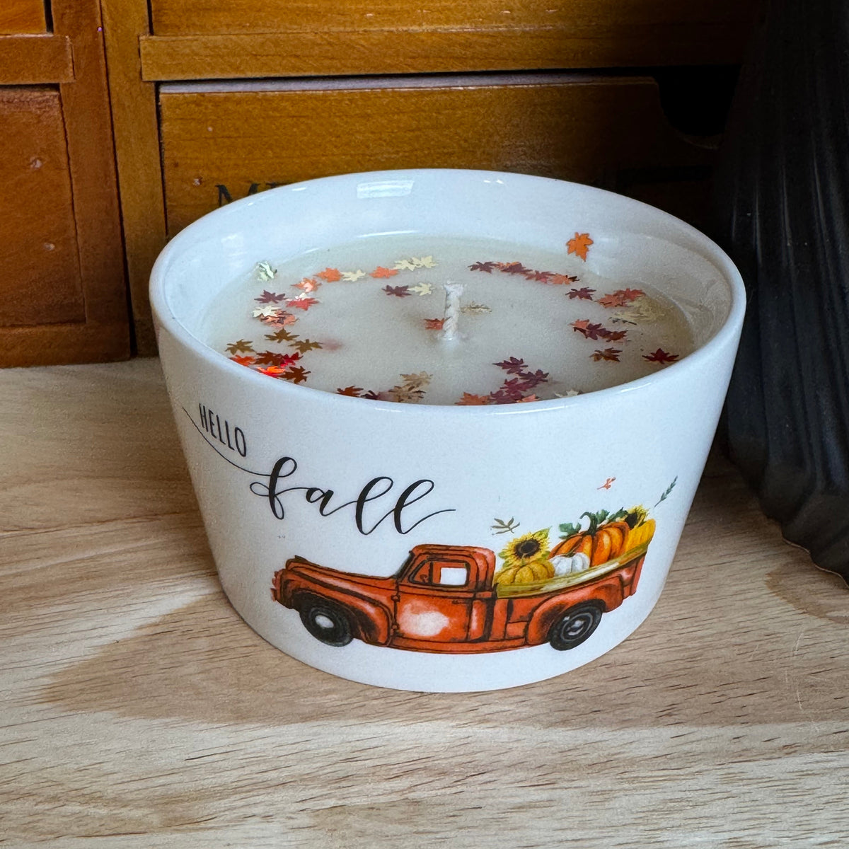A white ceramic candle jar with a 'Hello fall' autumn truck illustration, filled with wax and decorated with dried fall leaves, sitting on a wooden surface.