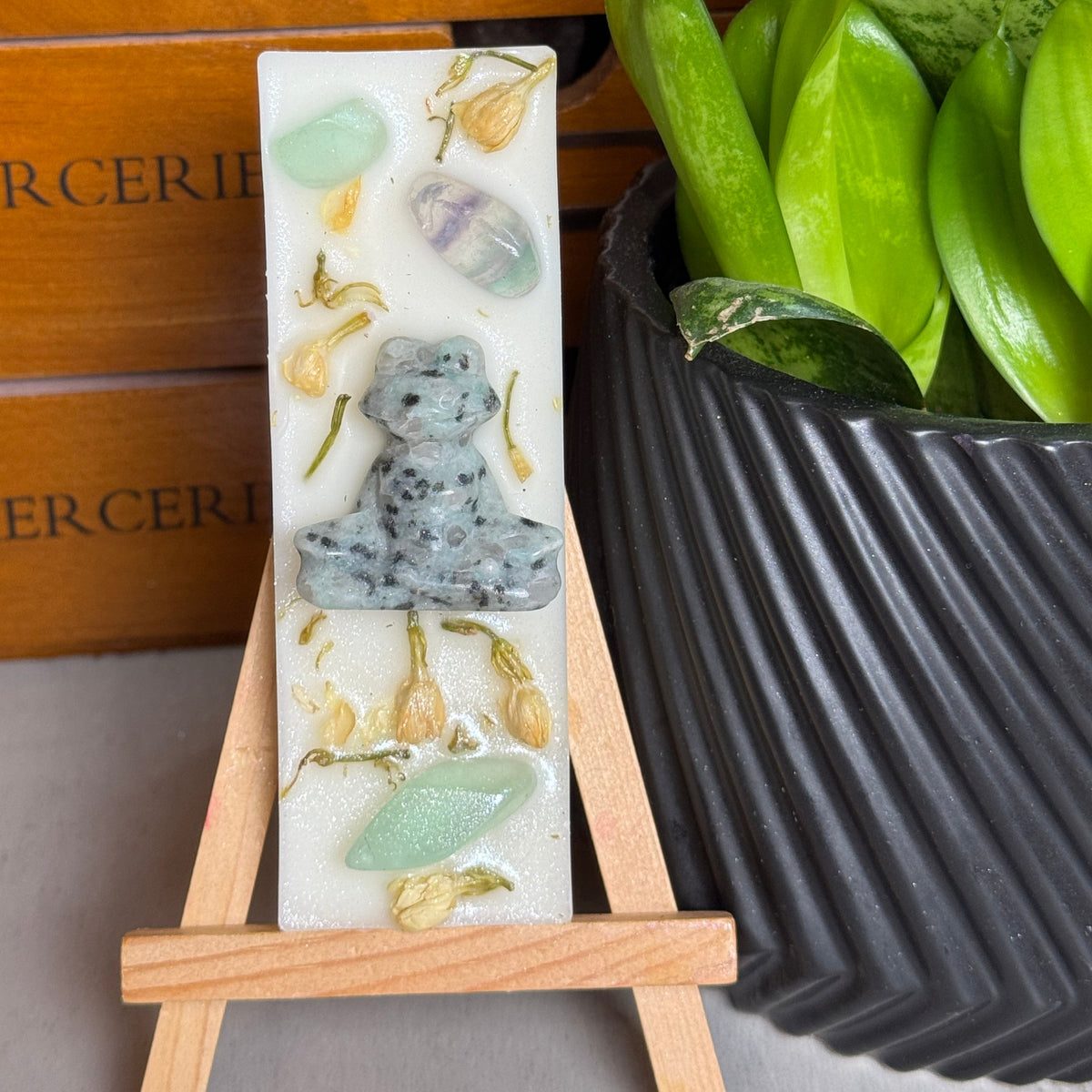 A rectangular white resin candle displayed on a small wooden easel, featuring a gray frog figure and green stone inclusions. The candle is styled in front of a wooden drawer and next to a potted green plant.