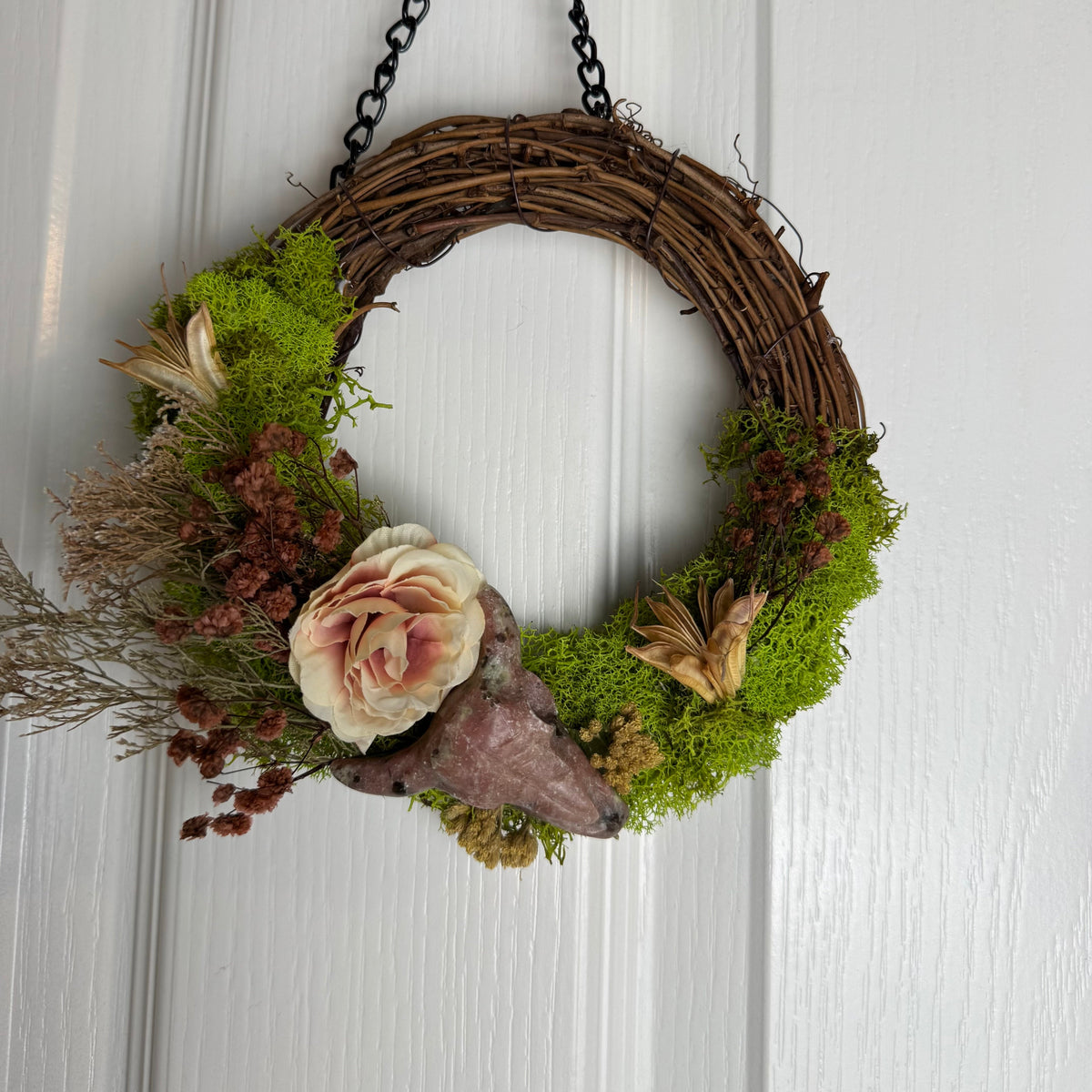 A rustic hanging wreath made of brown grapevine with bright green moss, dried pinkish-brown flowers, a pale cream rose, and a large natural shell, hanging on a white paneled door.