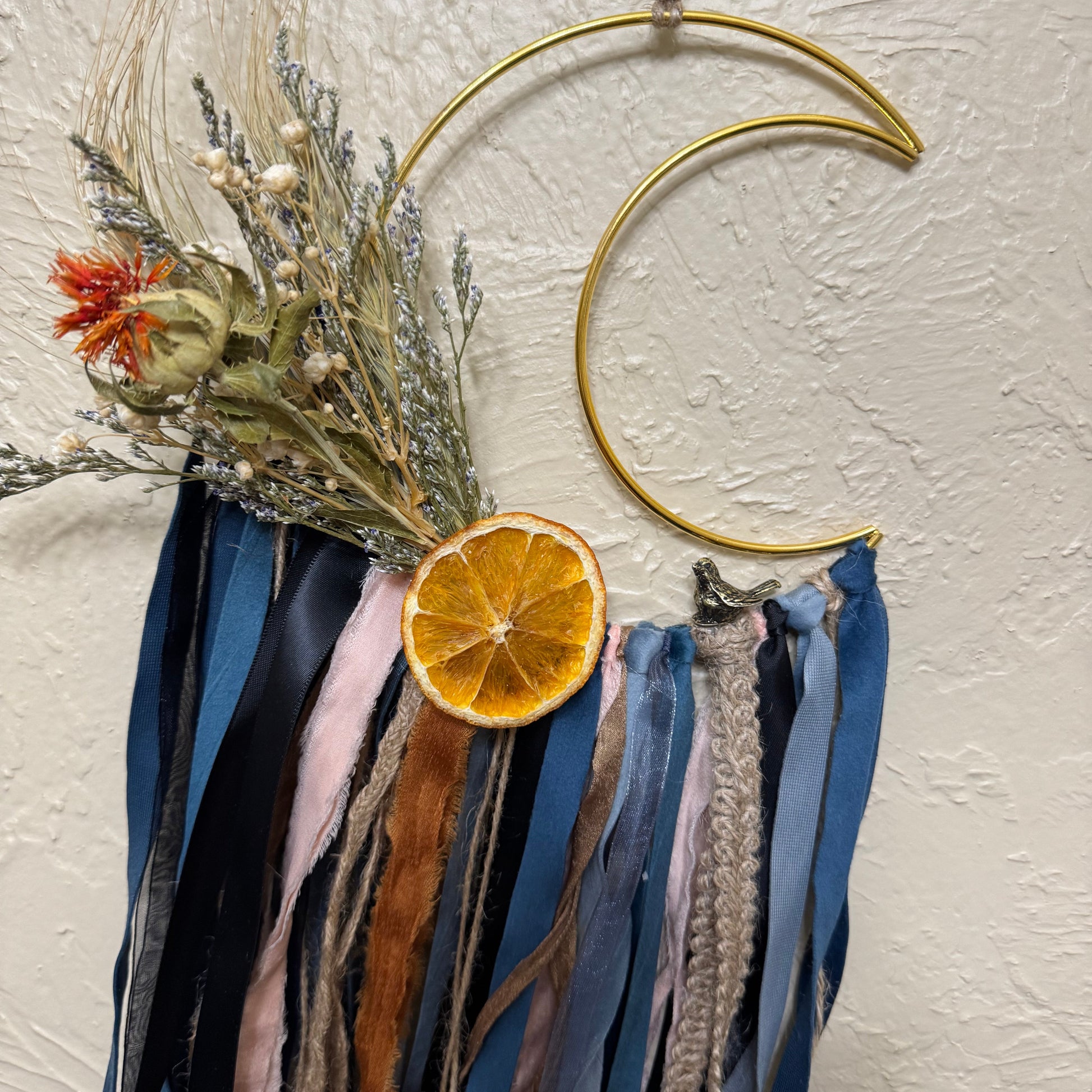 Decorative wall hanging with dried flowers, orange slice, and ribbons on a textured wall.