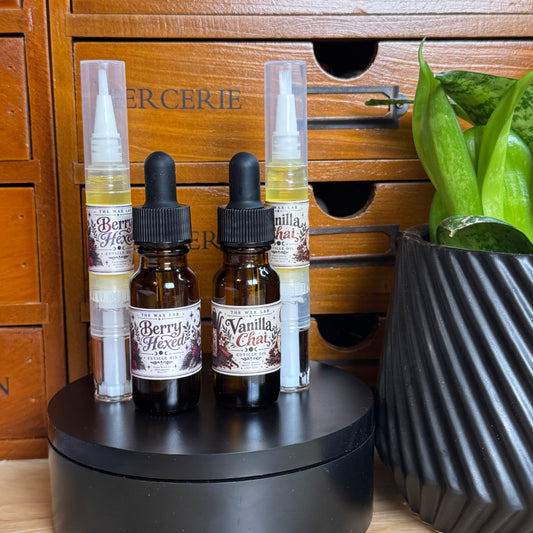 Four small amber dropper bottles and two clear spray bottles with white caps, labeled Berry & Vanilla, arranged on a round black display stand. The setup is placed on a wooden surface in front of a wooden drawer chest and next to a potted green plant.