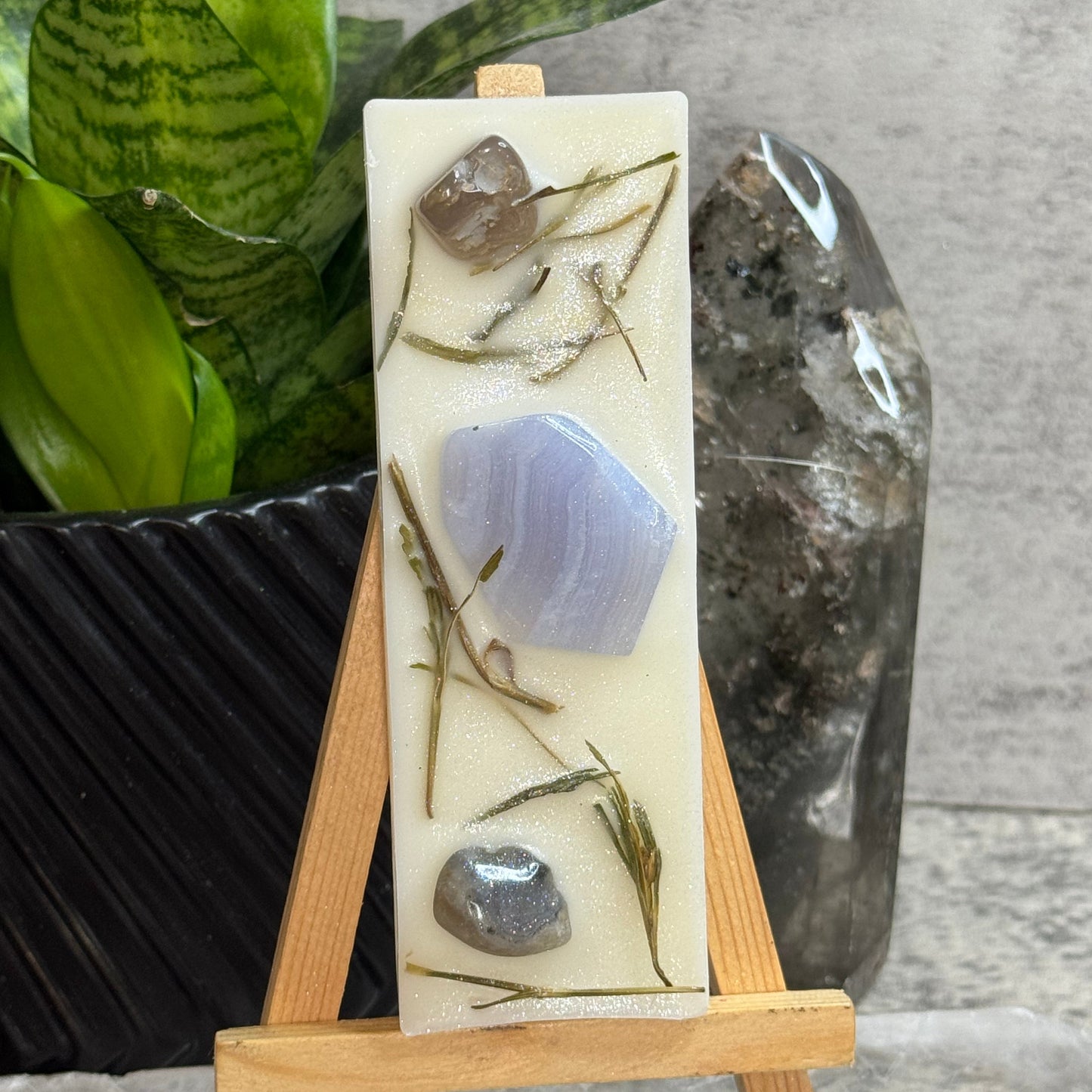 A rectangular cream-colored resin candle displayed on a small wooden easel. The candle is decorated with dried botanicals and polished stones, including a prominent lavender agate piece.