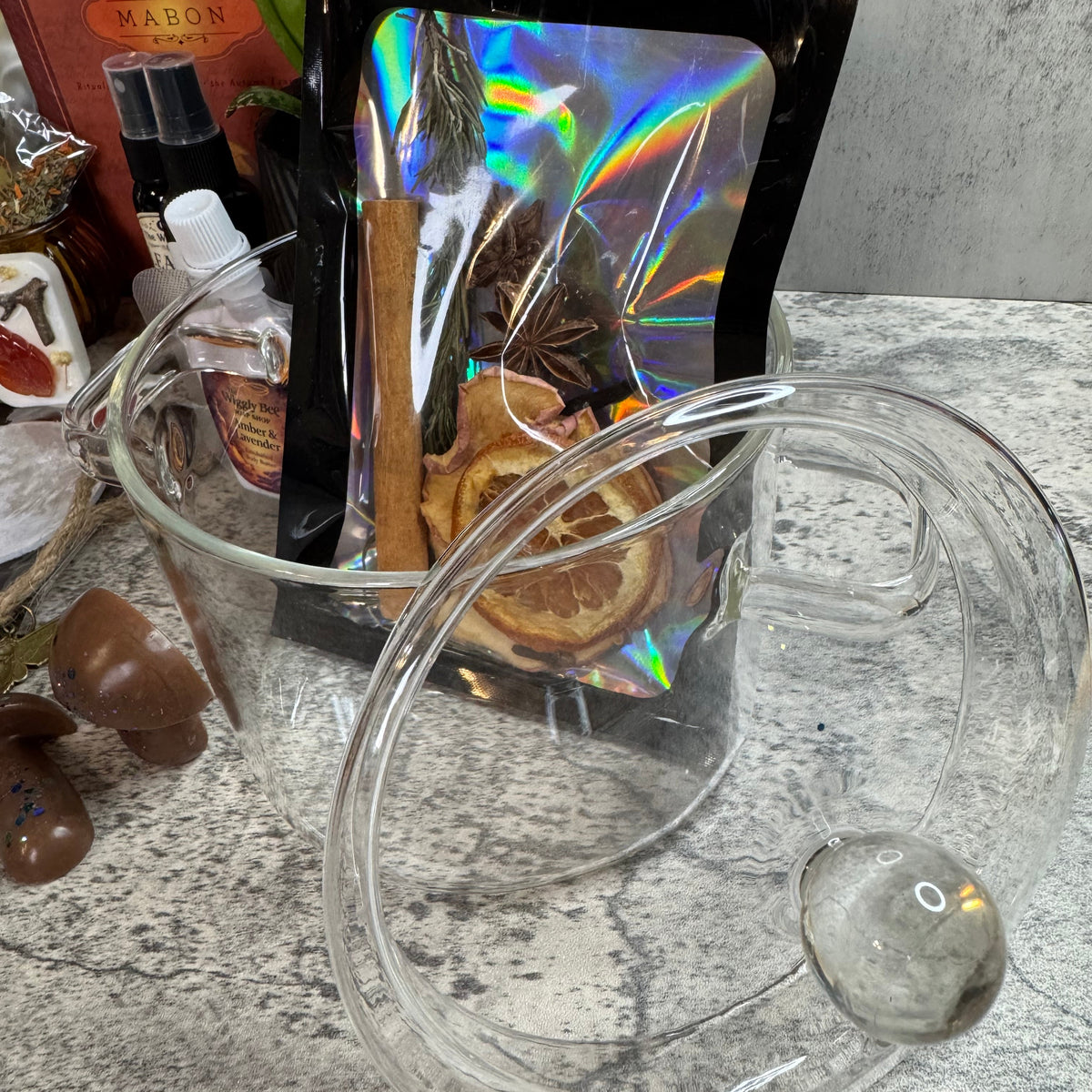 A clear glass mug with a handle and a matching clear lid, shown in front of a black resealable pouch with an iridescent window containing dried orange slices and cinnamon sticks, on a speckled gray countertop.