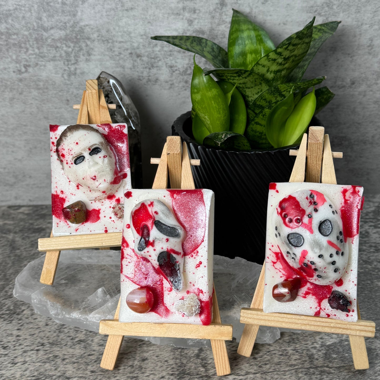 Three small canvas art prints featuring horror mask designs with red splatter details, displayed on miniature wooden easels against a gray textured wall with a green plant in the background.