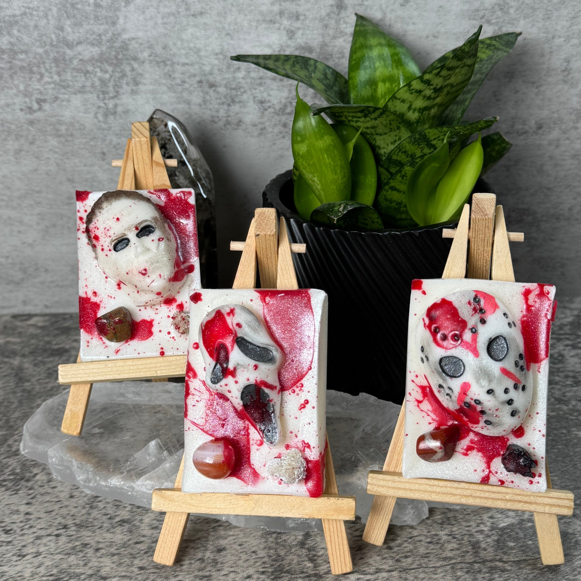 Three small canvas art prints featuring horror mask designs with red splatter details, displayed on miniature wooden easels against a gray textured wall with a green plant in the background.