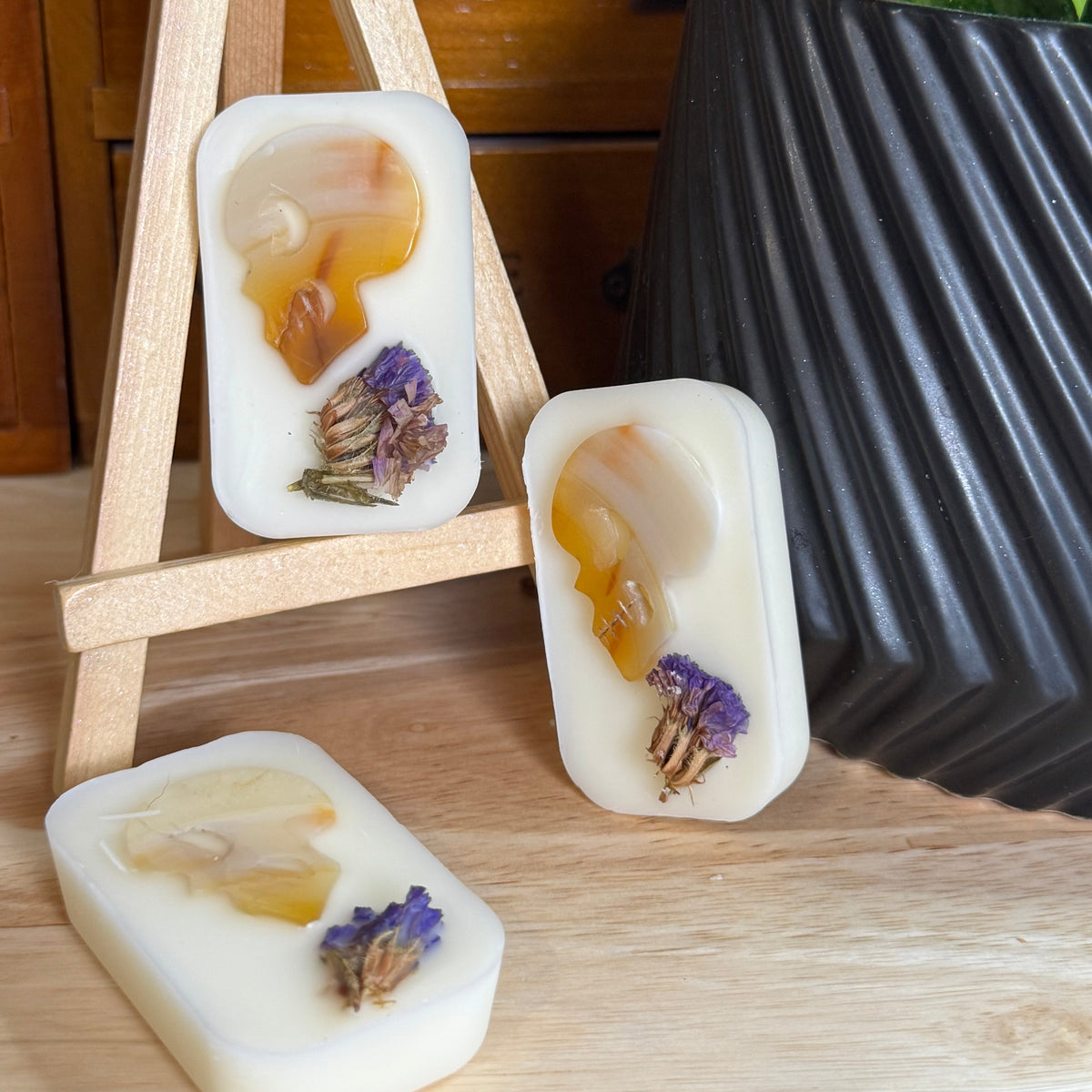 Three rectangular cream-colored handmade soaps with dried botanical inclusions, displayed on a wooden surface with a small wooden easel and a black ribbed planter in the background.