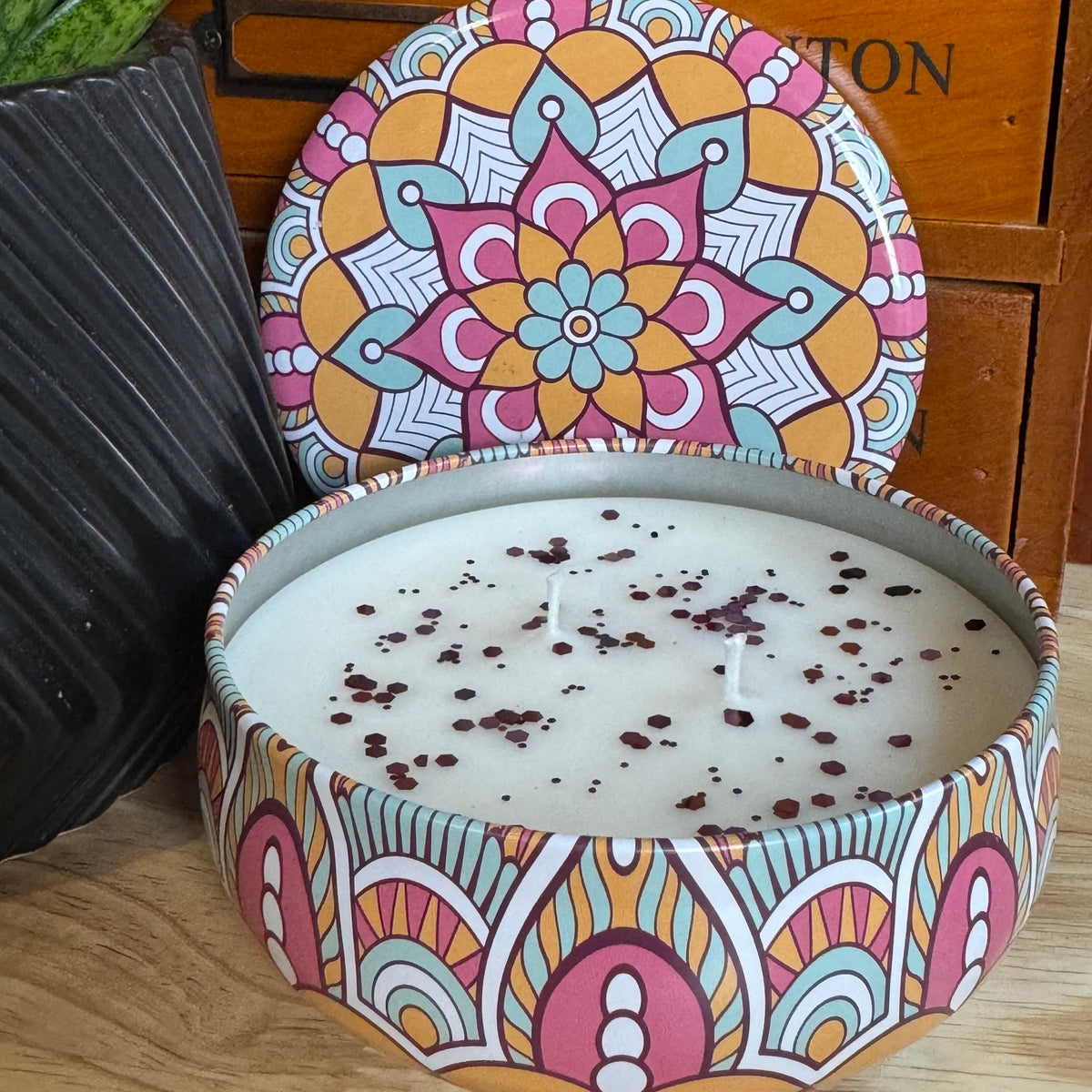 A round tin candle with white wax and dark speckles, shown open with its matching lid leaning behind it. The tin and lid feature a colorful mandala-style pattern in pink, orange, yellow, and blue.