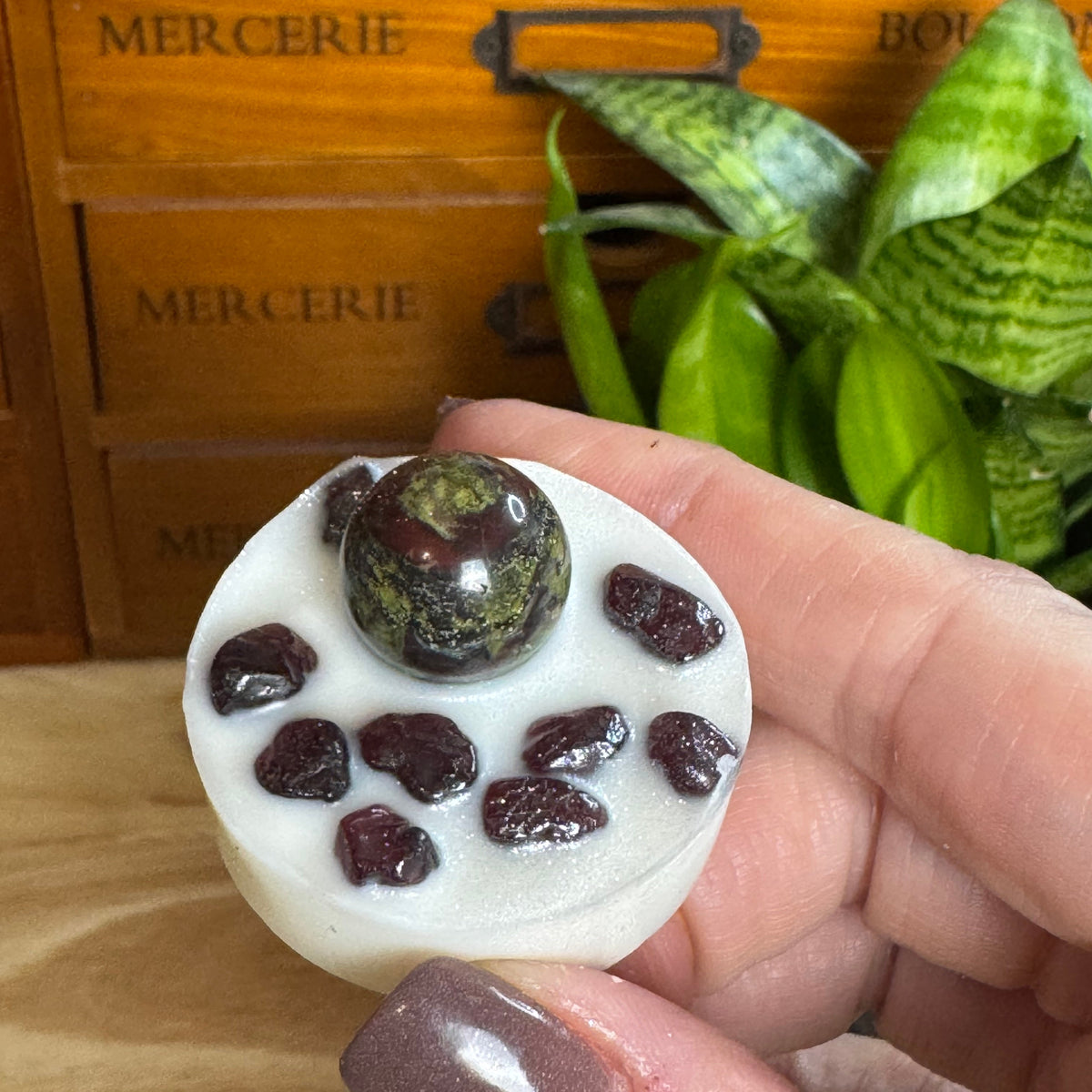 A hand holds a small white circular decorative object with a glossy dark green stone on top and several dark red beads around it, set against a wooden drawer and green plant background.