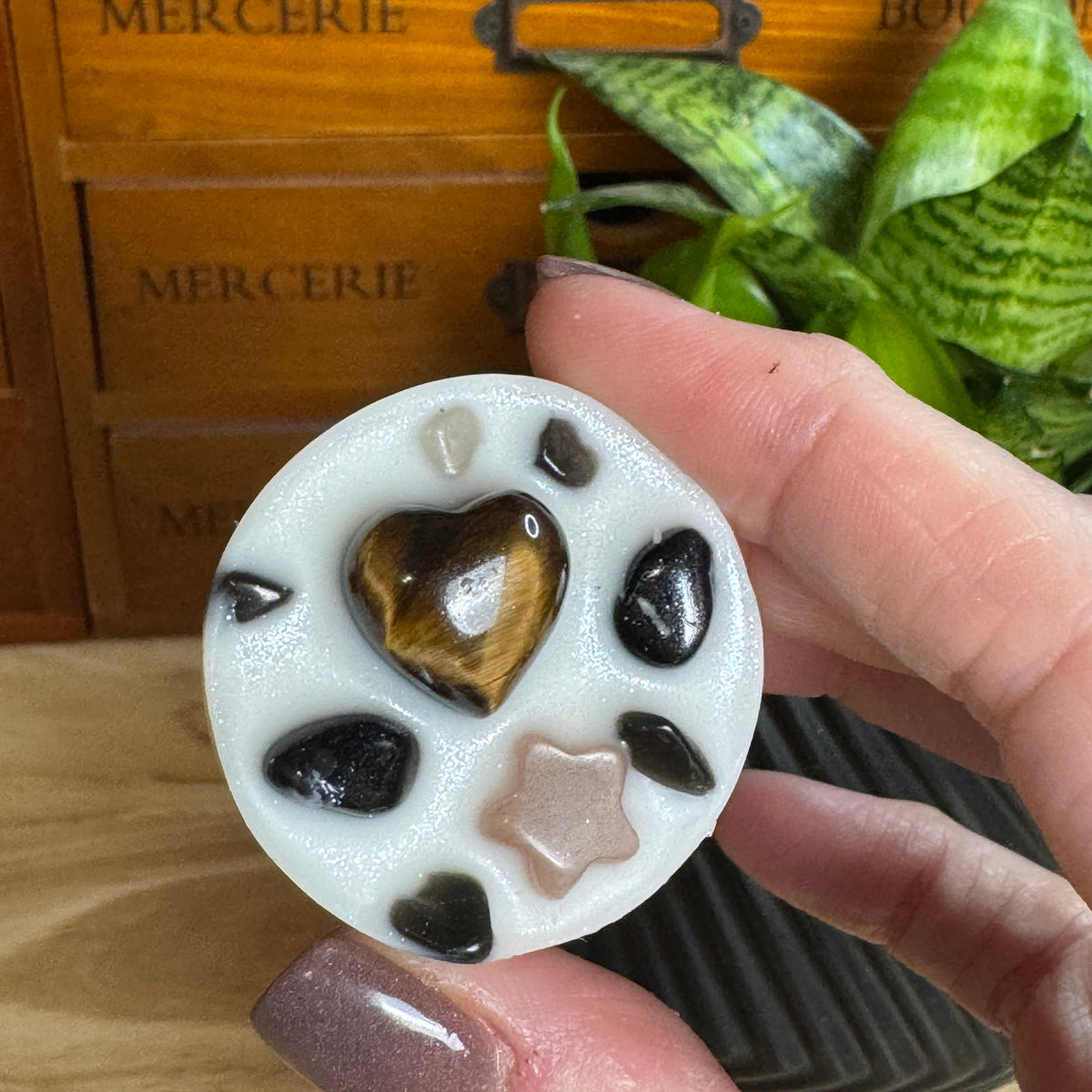 A hand holds a round white resin coaster or decorative pad featuring a tiger's eye heart, black stones, and a pale pink star, with a wooden drawer and green plant in the background.