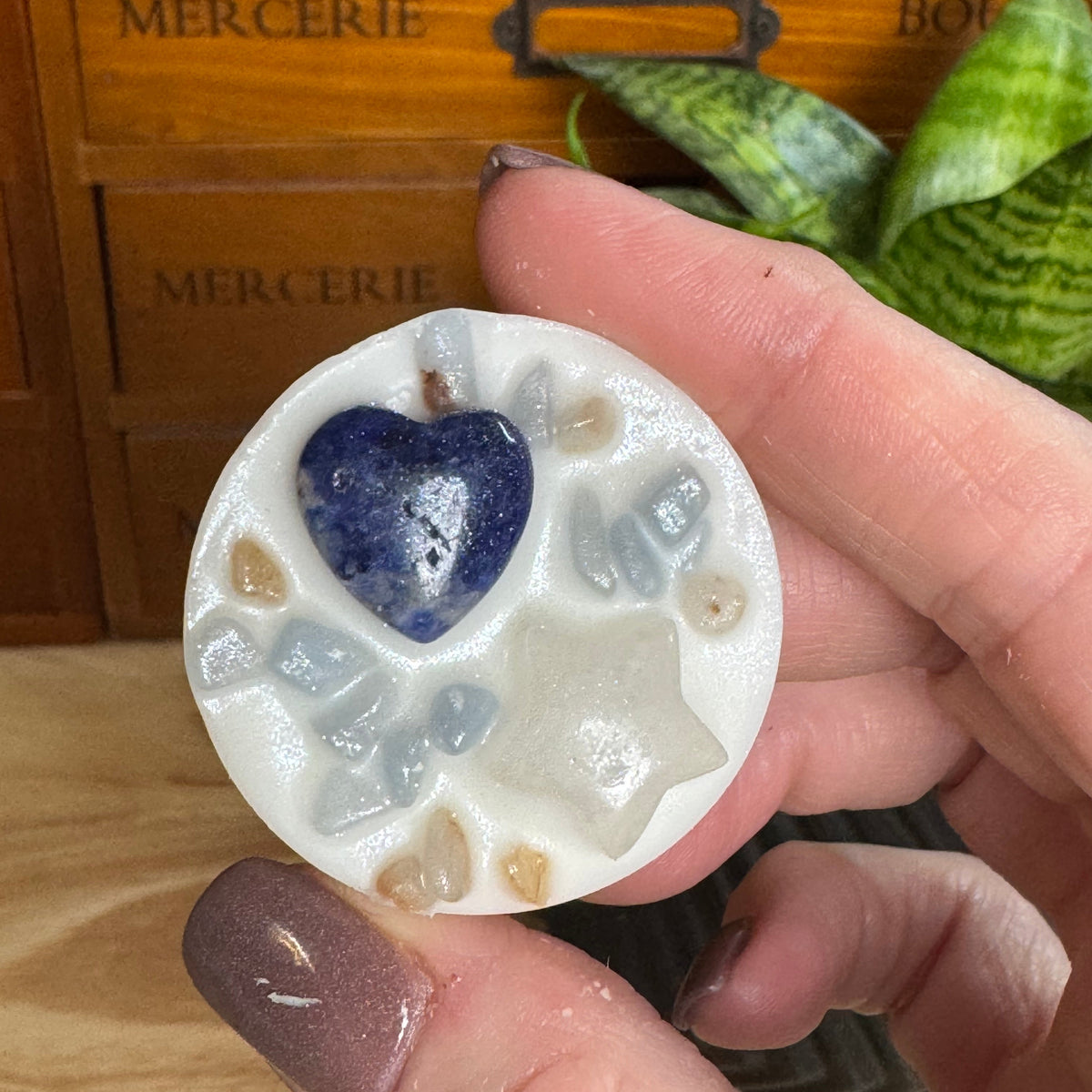 A hand holds a round white resin plaque featuring a polished blue heart and small clear and beige stones, with a wooden drawer and green plant in the background.