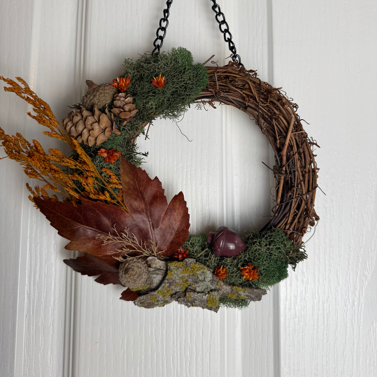 An autumn-themed grapevine wreath hanging on a white paneled door. The wreath is decorated with brown leaves, pinecones, green moss, orange dried flowers, acorns, and small red berries, suspended by a black metal chain.