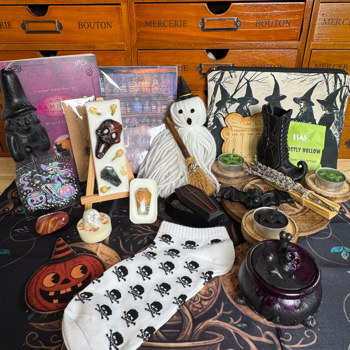 A styled flat-lay of a Halloween-themed product collection featuring a white crew sock with a black skull pattern, a white ghost figurine, a broom, a witch hat, a cauldron, a pumpkin, and other spooky accessories arranged on a dark patterned mat in front of wooden drawers labeled with shop names.