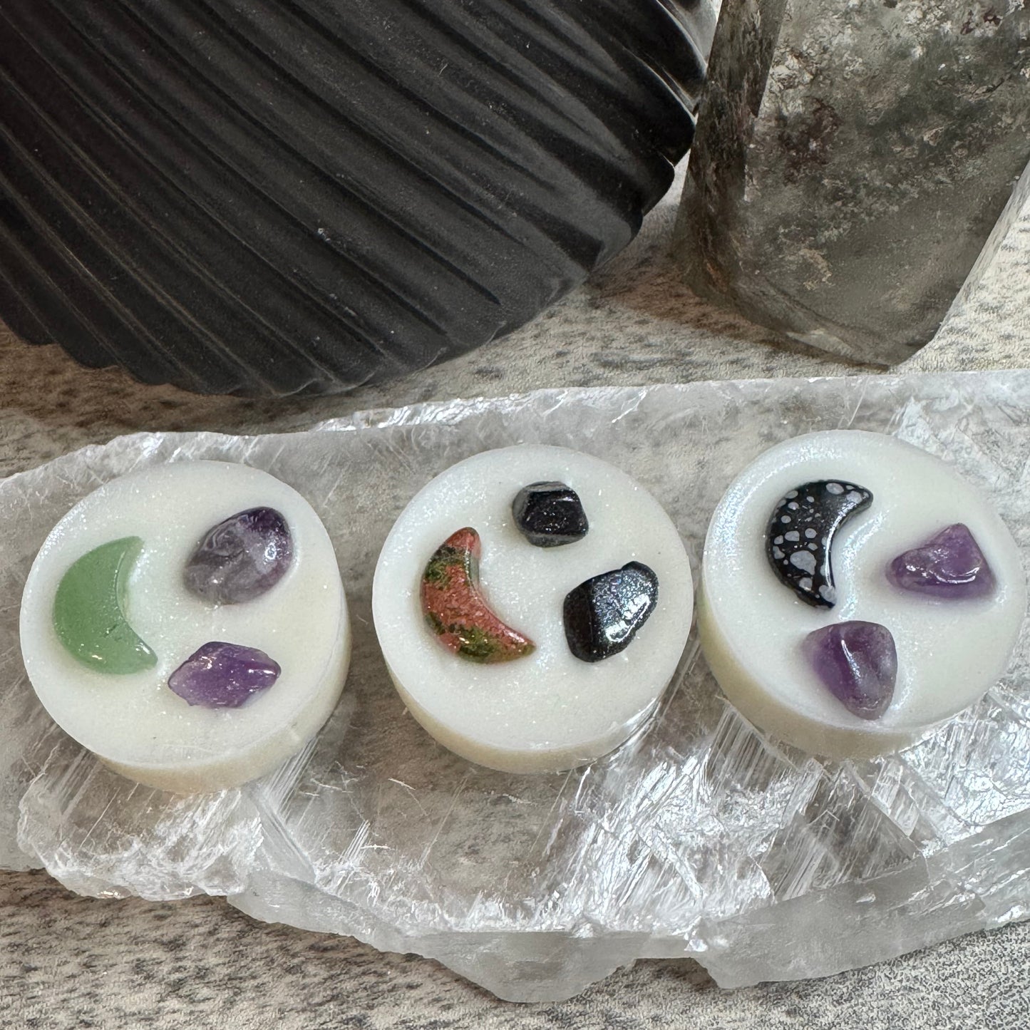 Three round white wax melts or soaps with embedded polished stones, arranged on a clear crystal sheet on a speckled stone surface. A black ribbed planter and a gray crystal tower are blurred in the background.