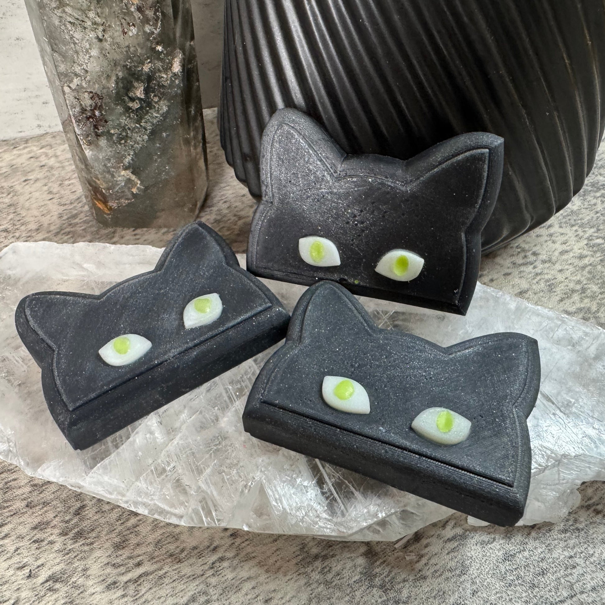 Three black cat-shaped soap bars with green eyes on a stone surface.