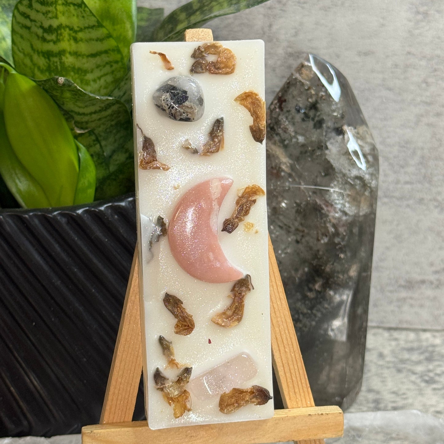 A rectangular cream-colored resin candle displayed on a small wooden easel. The candle features embedded crystals, including a prominent pink moon-shaped crystal, brown dried botanical pieces, and a clear quartz crystal.