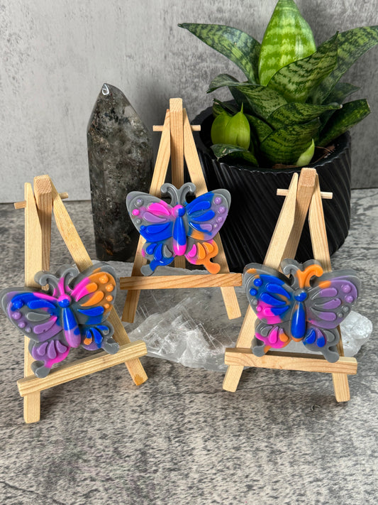 Colorful butterfly decorations on a wooden stand with a plant and black container in the background.
