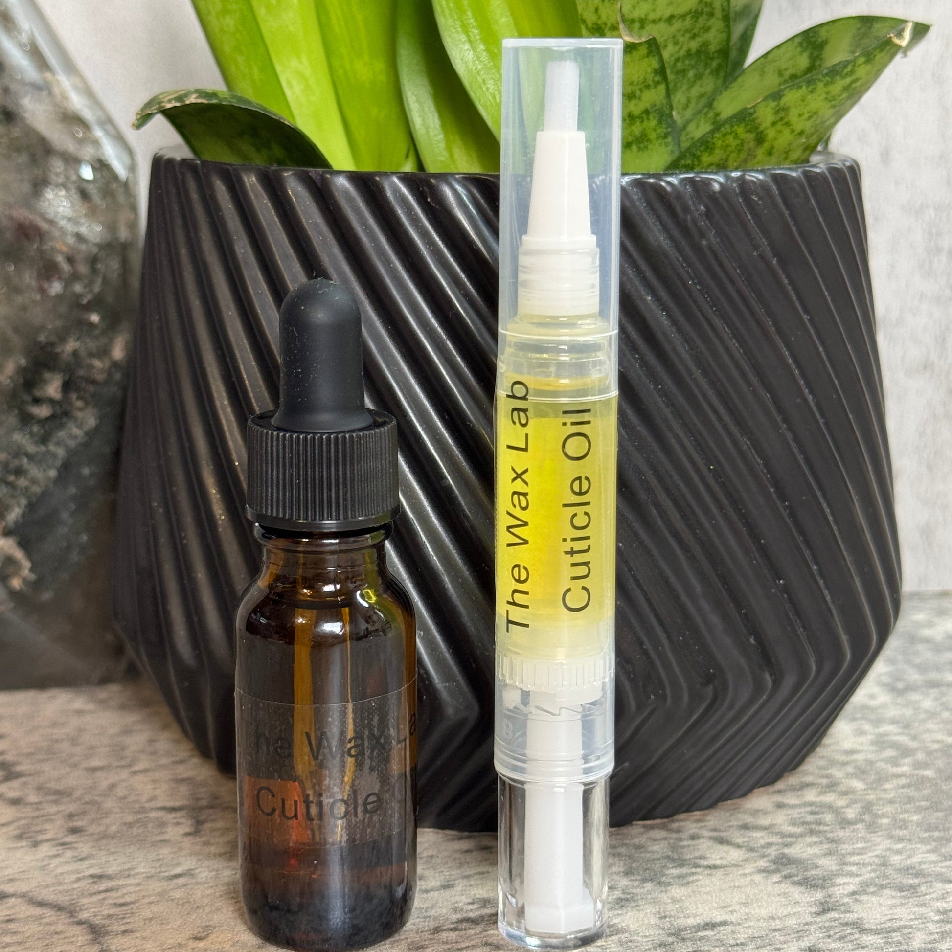 Two skincare products from The Wax Lab are displayed on a stone surface in front of a black textured planter with green snake plant leaves. The product on the left is a dark amber glass dropper bottle labeled 'Cuticle Oil', and the product on the right is a clear plastic tube containing pale yellow cuticle oil with a white applicator, labeled 'The Wax Lab Cuticle Oil'.