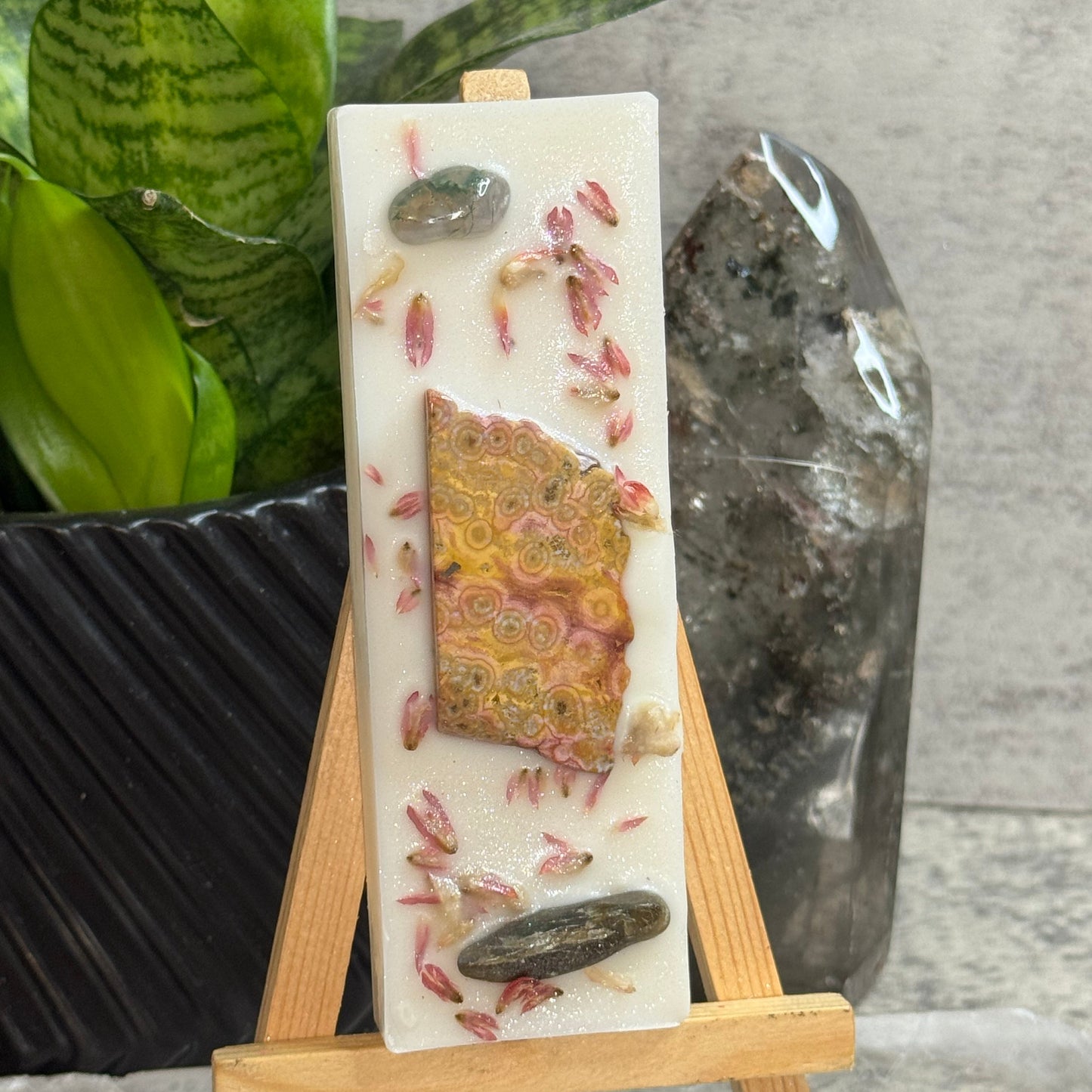 A rectangular white resin candle displayed on a small wooden easel. The candle features embedded natural elements including a piece of patterned agate, smooth river stones, and dried pink botanical pieces.