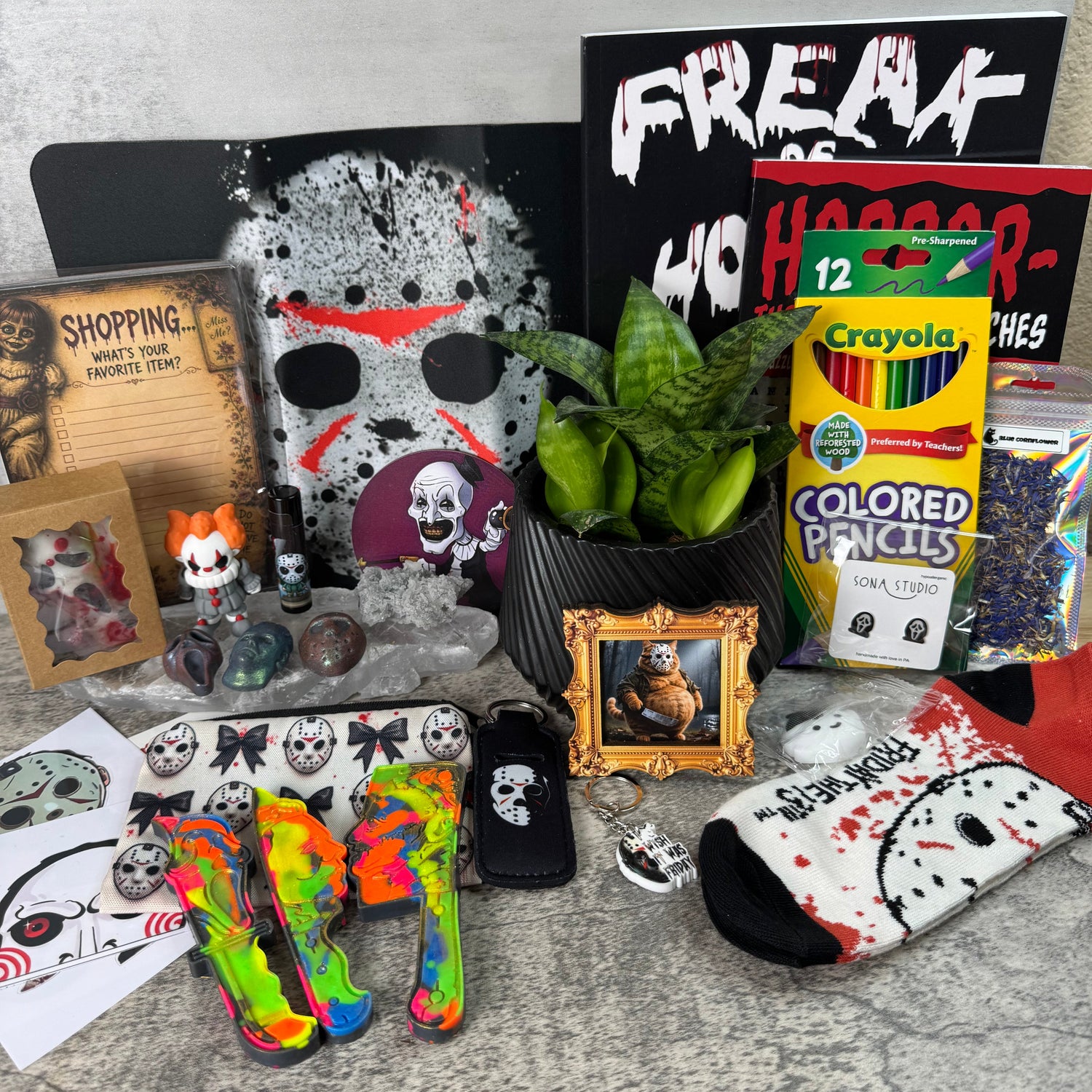 A Halloween-themed product display featuring a black and white horror mask, a black sign reading 'FREAKS WHO HATE', a box of Crayola colored pencils, a small framed picture, a potted plant, a small figurine, stickers, a neon rainbow high heel, and a black and white sock with a skull pattern, arranged on a gray stone-textured surface.