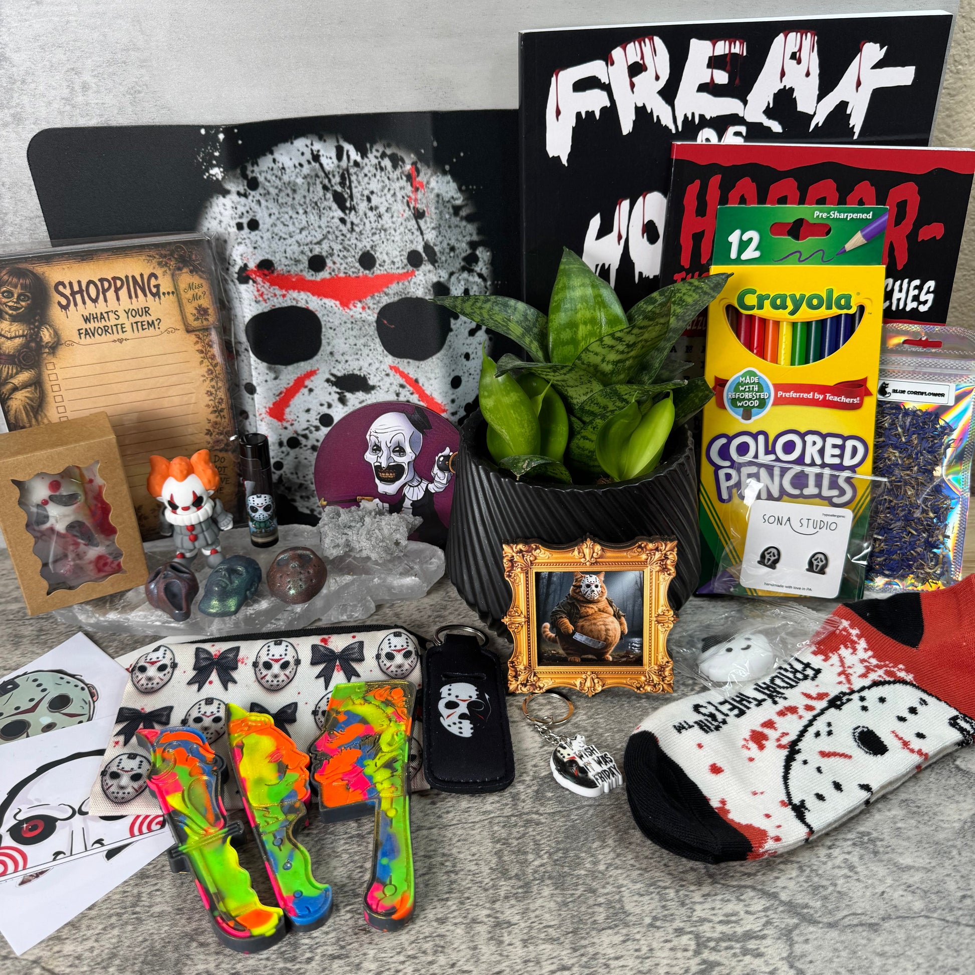 A Halloween-themed product display featuring a black and white horror mask, a black sign reading 'FREAKS WHO HATE', a box of Crayola colored pencils, a small framed picture, a potted plant, a small figurine, stickers, a neon rainbow high heel, and a black and white sock with a skull pattern, arranged on a gray stone-textured surface.