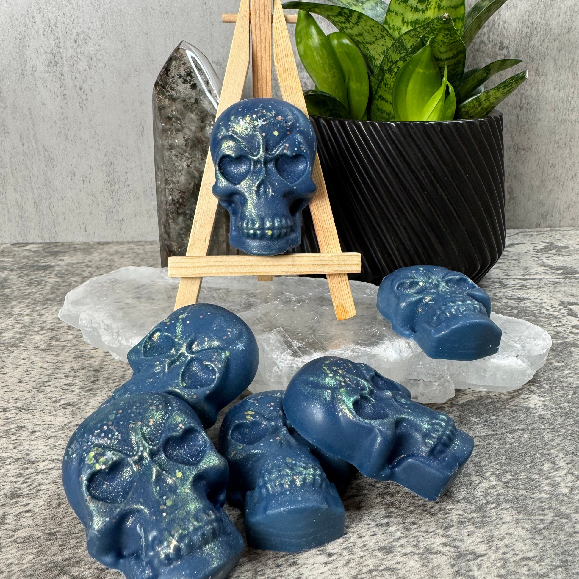 Blue skull-shaped soap bars on a stand with a black bar of soap and green leaves on a gray surface