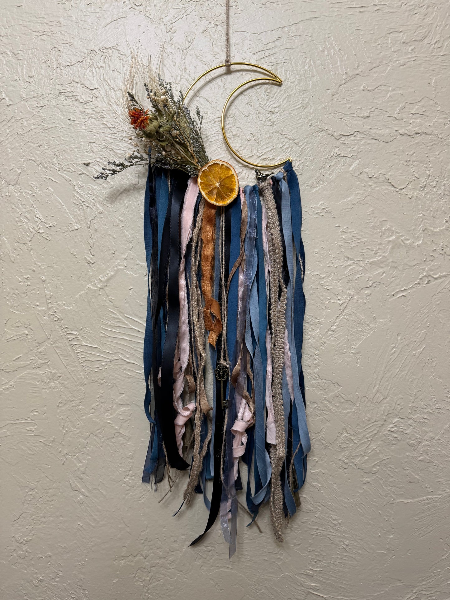 Decorative wall hanging with ribbons and a gold crescent moon on a light gray wall.