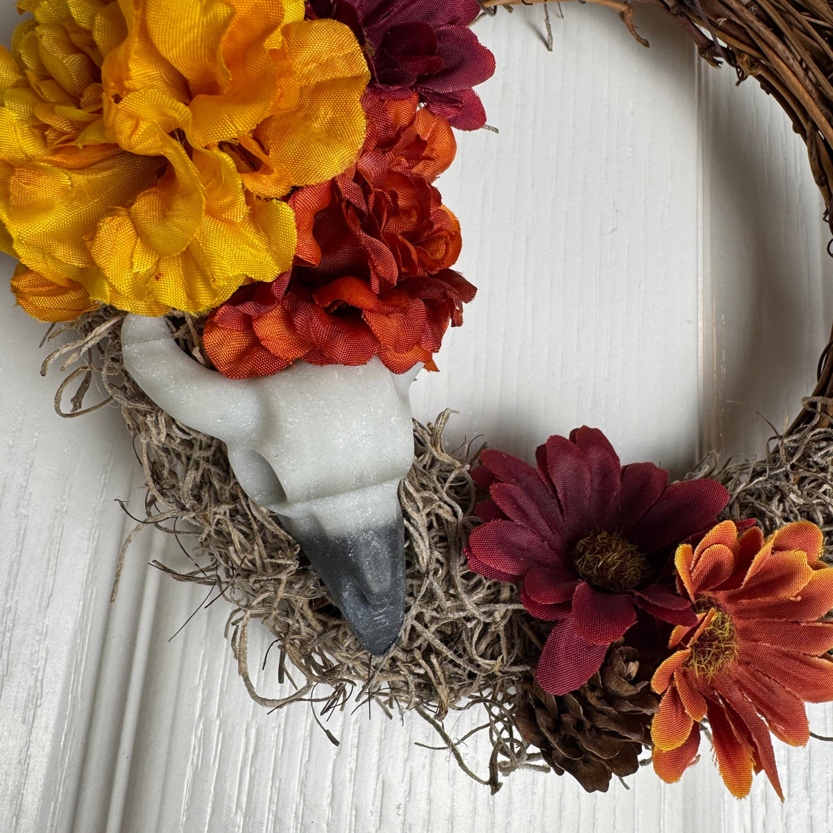 A rustic autumn-themed wreath made of brown grapevine branches, decorated with large yellow, orange, and burgundy fabric flowers, a white ostrich feather, and a pinecone, hanging against a white paneled wall.