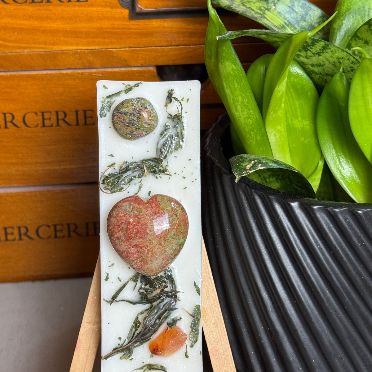 A rectangular white resin candle displayed on a small wooden easel, featuring embedded botanical elements and a large heart-shaped crystal. The candle is styled in front of a wooden drawer and next to a potted green plant.