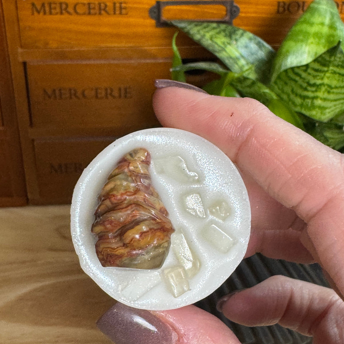 A hand with mauve glitter nail polish holds a small oval white resin tray containing a marbled brown and gold crescent moon charm and clear crystal pieces. The background shows a wooden drawer with 'MERCERIE' text and a green snake plant.