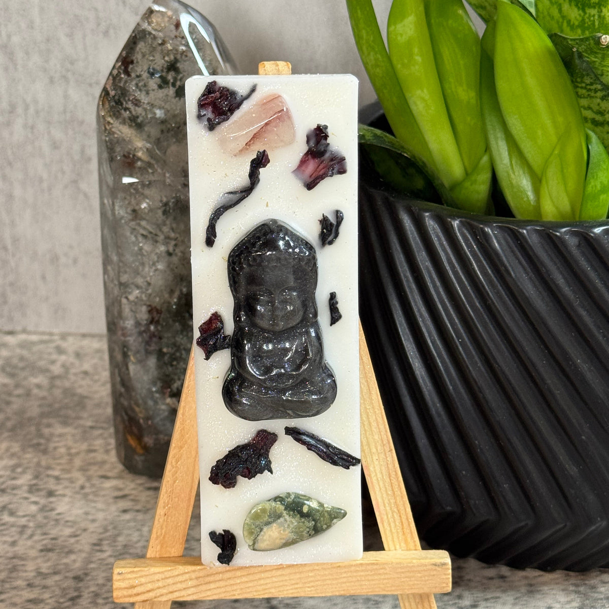 A vertical white resin candle displayed on a small wooden easel, featuring a carved black Buddha figure and embedded crystals, with a plant and crystal cluster in the background.