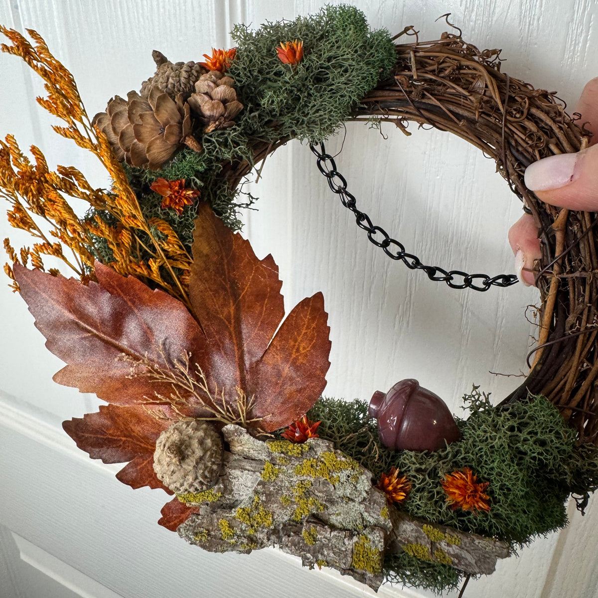 An autumn-themed grapevine wreath decorated with dried brown leaves, pinecones, green moss, orange flowers, and a small acorn, being held on a white paneled door by a hand.