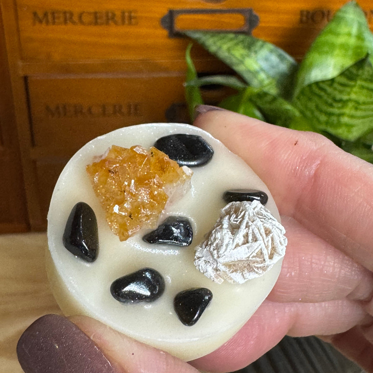 A hand holds a round cream-colored soap or candle featuring black polished stones, a textured orange crystal, and a white woven fiber accent, with a wooden drawer and green plant in the background.