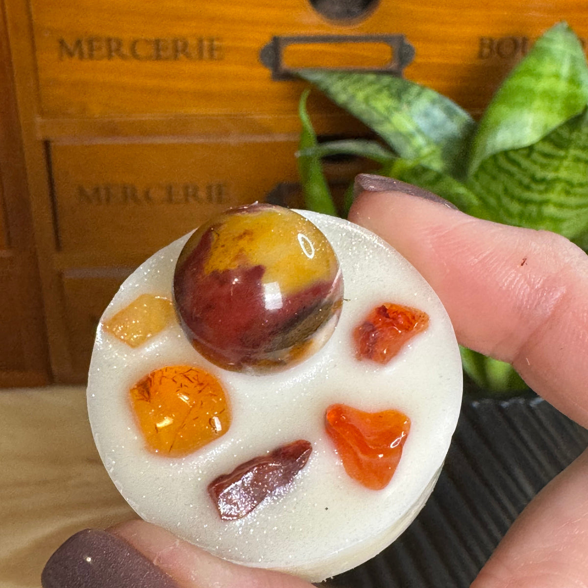 A hand with mauve nail polish holds a round white candle topped with six polished gemstone pieces in amber, orange, and reddish-brown tones. A wooden cabinet with 'MERCERIE' text and a green plant are blurred in the background.