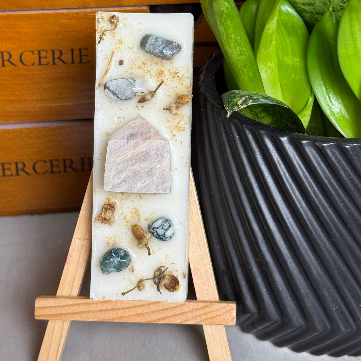 A rectangular handmade soap bar with embedded stones and dried botanicals, displayed on a small wooden easel stand. The background features a wooden drawer with 'MERCERIE' text and a green potted plant.