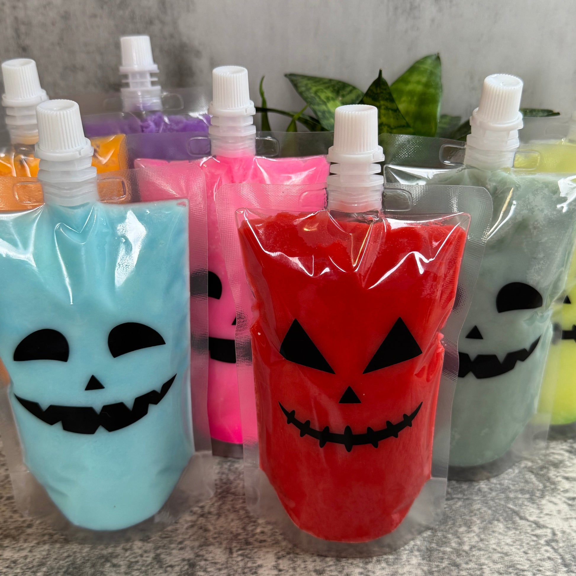 A group of Halloween-themed squeeze pouches filled with colorful slime, decorated with jack-o'-lantern faces. The front pouches are bright red and light blue, with pink, green, purple, and yellow pouches lined up behind them on a speckled gray countertop.