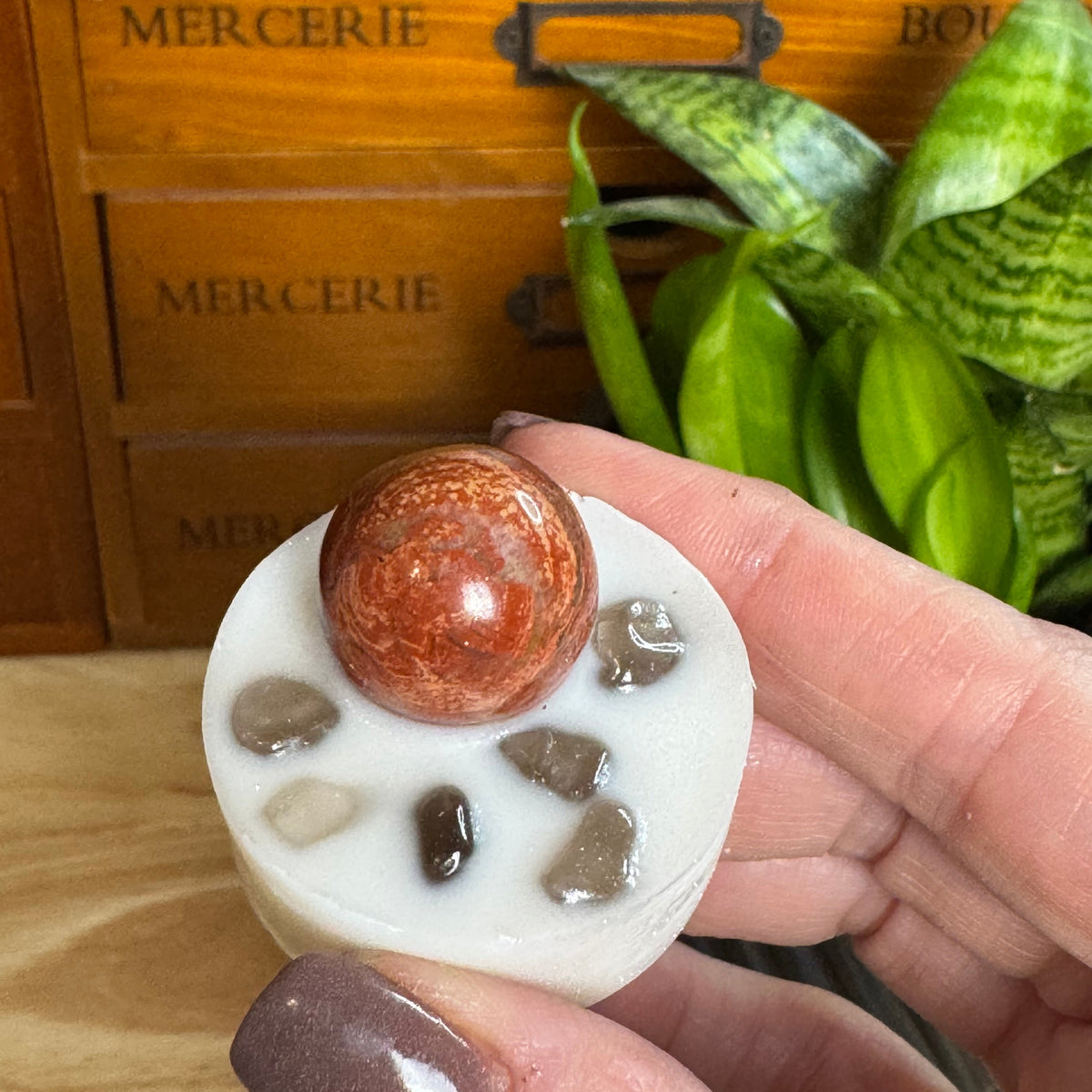 A hand with mauve nail polish holds a small white resin candle featuring a round orange-brown stone on top and several small polished stones embedded in the wax. The background shows a wooden drawer unit with the word 'MERCERIE' and a green plant.