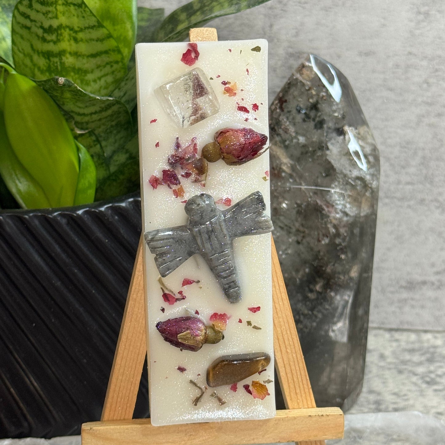 A rectangular white resin candle displayed on a small wooden easel. The candle contains embedded dried rosebuds, a gray stone airplane, a clear crystal cube, and a small amber piece.