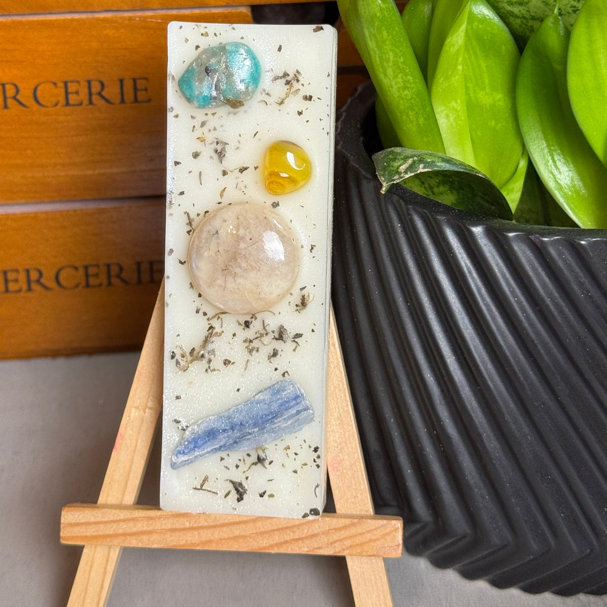 A rectangular white resin candle displayed on a small wooden easel, featuring embedded polished stones in blue, yellow, and peach, with dried botanical flecks. The candle is styled in front of a wooden drawer and next to a potted green plant.