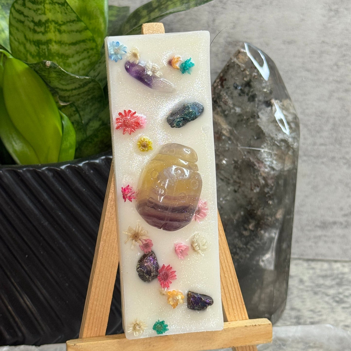 A rectangular white resin candle displayed on a small wooden easel. The candle features embedded polished stones in purple, amber, green, and pink, along with tiny dried flower petals.