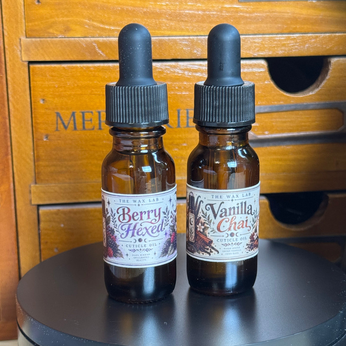 Two amber glass dropper bottles with black caps and white labels, standing on a round black platform in front of a wooden drawer unit. The left label reads 'Berry Hexed' and the right label reads 'Vanilla Chat'.