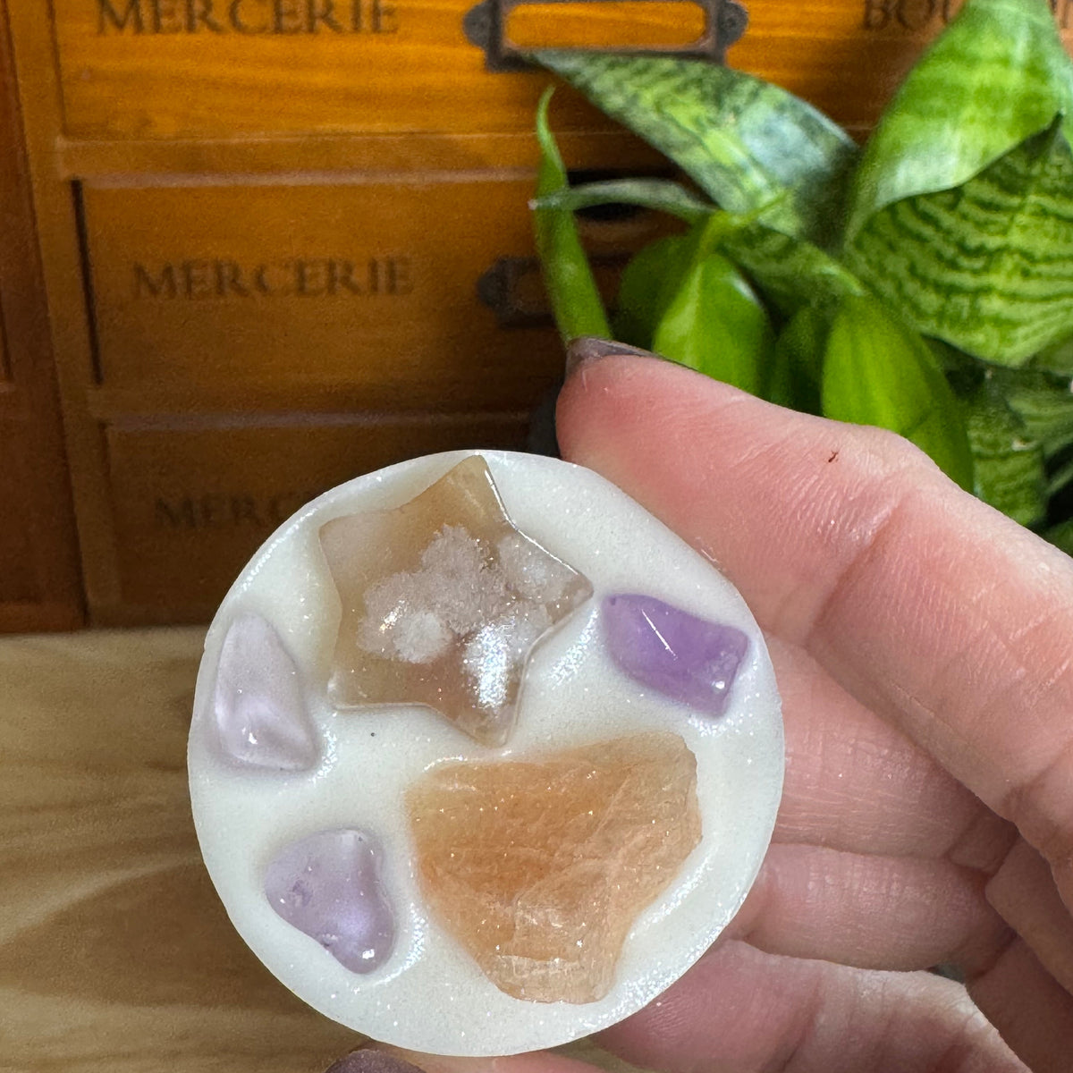 A hand holds a round white wax melt or candle tin filled with natural crystals, including a star-shaped crystal, rose quartz, and purple amethyst, with a wooden drawer and green plant in the background.