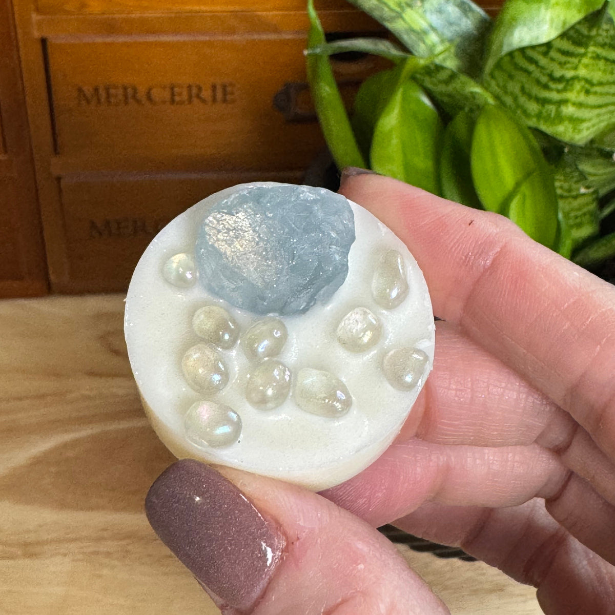 A hand holds a round white soap bar decorated with a blue-gray stone and small clear pearls, set against a background of a wooden drawer and green plants.