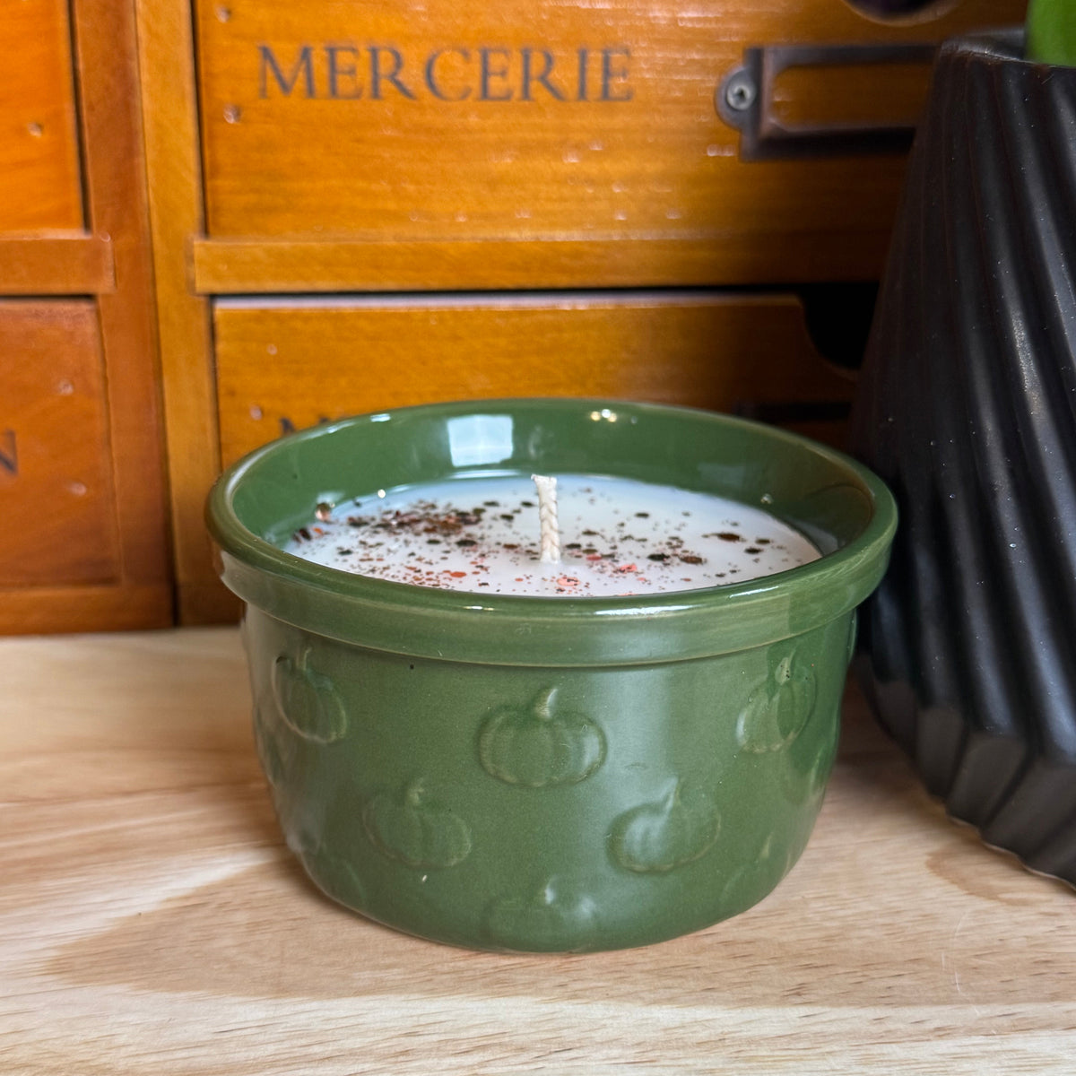A green ceramic candle jar with a pumpkin embossed pattern sits on a wooden table in front of a wooden drawer cabinet with the word 'MERCERIE' on it.